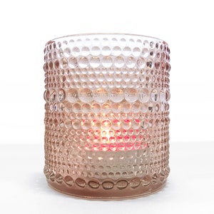 May include: A pink glass votive candle holder with a textured, bubble-like pattern. The candle holder is cylindrical and contains a lit candle, casting a warm glow. The overall aesthetic is elegant and decorative.