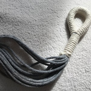 May include: Close-up of a rope with a loop. The loop is made of thick, tightly wound white rope. The other end of the rope is a bundle of gray strands. The background is a textured, off-white surface.