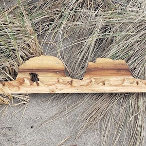 May include: A wooden wall art piece depicting a stylized mountain range with a wave pattern at the base. The piece is made of light brown wood and has a natural finish.