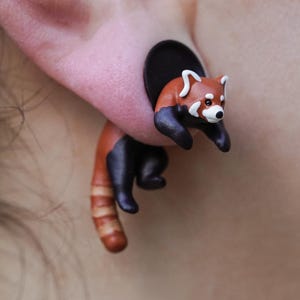 May include: A red panda charm earring with a black and brown body and a white face. The earring is made of polymer clay and is attached to a black post.