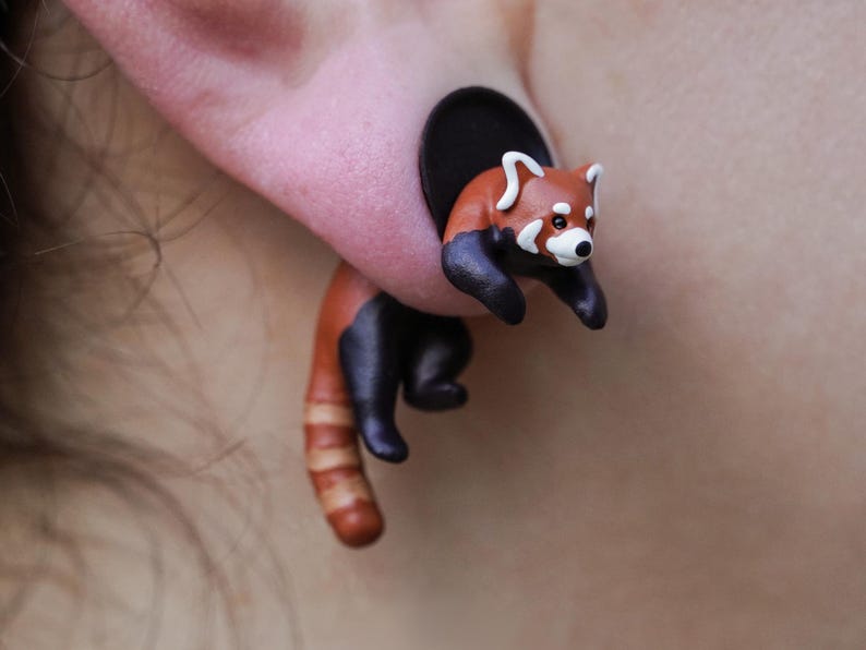 May include: A red panda charm earring with a black and brown body and a white face. The earring is made of polymer clay and is attached to a black post.