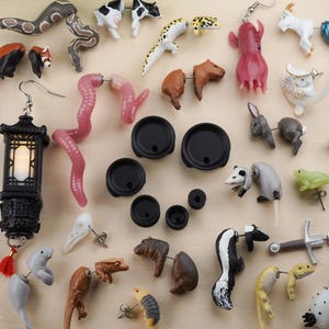 May include: A collection of colorful and whimsical earrings featuring various animal and nature-inspired designs. The earrings are made of different materials, including plastic, metal, and wood. Some of the designs include a red panda, a snake, a frog, a skunk, a seahorse, a jellyfish, and a lantern.