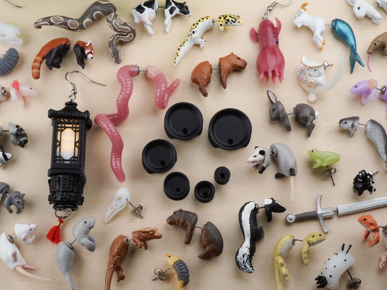 May include: A collection of colorful and whimsical earrings featuring various animal and nature-inspired designs. The earrings are made of different materials, including plastic, metal, and wood. Some of the designs include a red panda, a snake, a frog, a skunk, a seahorse, a jellyfish, and a lantern.