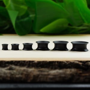 May include: A set of six black wooden ear gauges of varying sizes. The gauges are arranged in order of size, with the smallest gauge on the left and the largest gauge on the right.