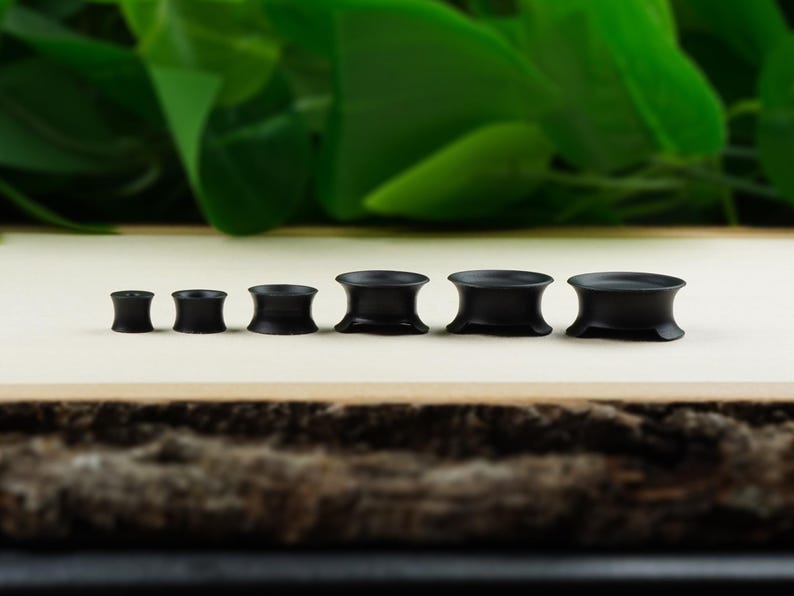 May include: A set of six black wooden ear gauges of varying sizes. The gauges are arranged in order of size, with the smallest gauge on the left and the largest gauge on the right.