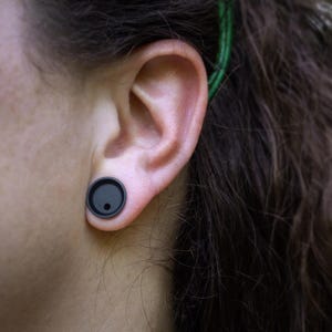 May include: A person with long dark hair wearing a black ear gauge in their left ear.