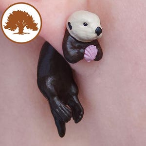 Op de afbeelding: Oorringen in de vorm van een otter. Eén oorring heeft een zwart otterlichaam en de andere een witgekopte otterkop met een roze schelp. De oorbellen staan tegen een lichtroze achtergrond. Een cirkelvormig logo met een boom staat in de linkerbovenhoek.