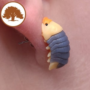 Puede incluir: Un pendiente de arcilla polimérica azul y naranja con forma de cochinilla. El pendiente está sujeto a un poste plateado.