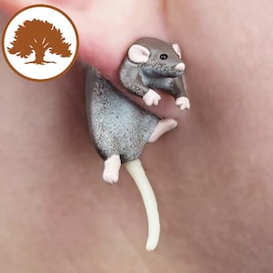Puede incluir: Un pequeño pendiente de ratón gris con una larga cola blanca. El ratón está posado en el lóbulo de la oreja de una persona.