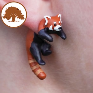 May include: Red panda dangle earrings made of polymer clay. The red panda is in a climbing pose with its tail curled around its body. The earrings are a reddish-brown color with black accents.