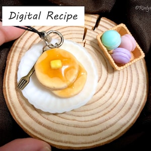 Könnte beinhalten: Ein Miniatur-Pfannkuchen-Schlüsselanhänger mit einem weißen Teller, einer Gabel und einem kleinen Stück Butter darauf. Der Schlüsselanhänger befindet sich auf einer Holzbasis mit einem kleinen Korb mit pastellfarbenen Macarons.