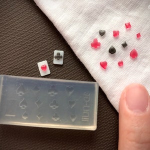 Könnte beinhalten: Eine transparente Silikonform mit 10 verschiedenen Formen von Spielkartenmotiven. Die Form befindet sich auf einer braunen Oberfläche. Auf einer weißen Oberfläche befinden sich außerdem mehrere kleine rote und schwarze Spielkartenmotive.