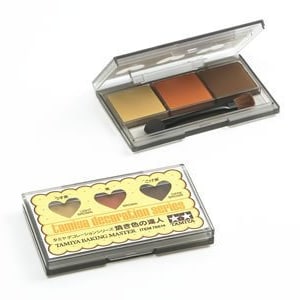 Op de afbeelding: Een Tamiya Baking Master set met drie acrylverven in een transparante kunststof doos met een klein penseel. De kleuren zijn lichtgeel, oranje en bruin. De doos heeft een label met de tekst "Tamiya Baking Master" en "Lining Decoration Set".