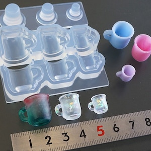 May include: A silicone mold for making miniature cups and mugs. The mold has eight cavities, each shaped like a different type of cup. There are also several small cups made from the mold, including a clear cup with a glittery design, a blue cup, a pink cup, and a clear cup with a green and red design.