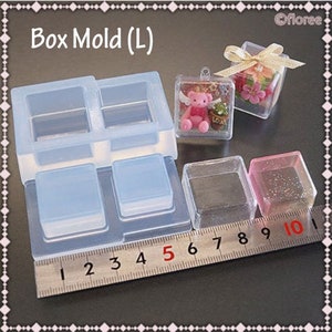 May include: A clear silicone mould for making small square boxes. The mould has four cavities, each measuring approximately 2.5 cm by 2.5 cm. The mould is shown next to a ruler, with the numbers 1 through 10 visible. The mould is also shown next to two small square boxes, one clear and one with glitter.