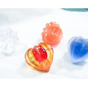 May include: Four glass paperweights in different shapes and colours. One is a clear flower shape, one is a red heart shape, one is a blue heart shape, and one is an orange heart shape with red and yellow swirls.