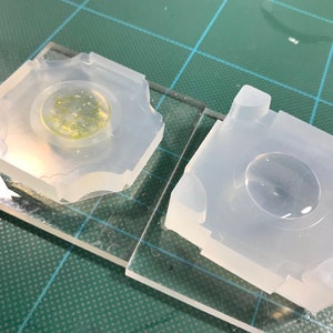May include: Two clear silicone molds for resin casting, one with a cured resin piece inside. The molds are square with a round indentation in the center.