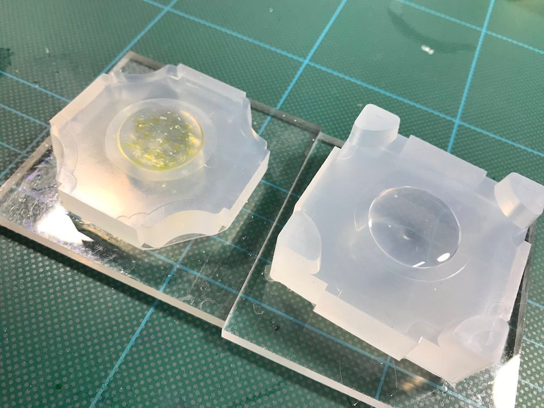 May include: Two clear silicone molds for resin casting, one with a cured resin piece inside. The molds are square with a round indentation in the center.