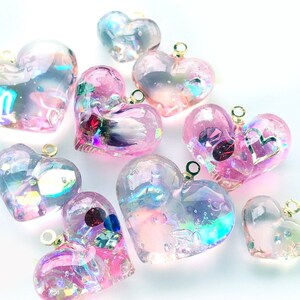 May include: A collection of iridescent pink heart-shaped resin charms with gold jump rings. The charms are filled with glitter and other embellishments, creating a sparkly and whimsical effect.