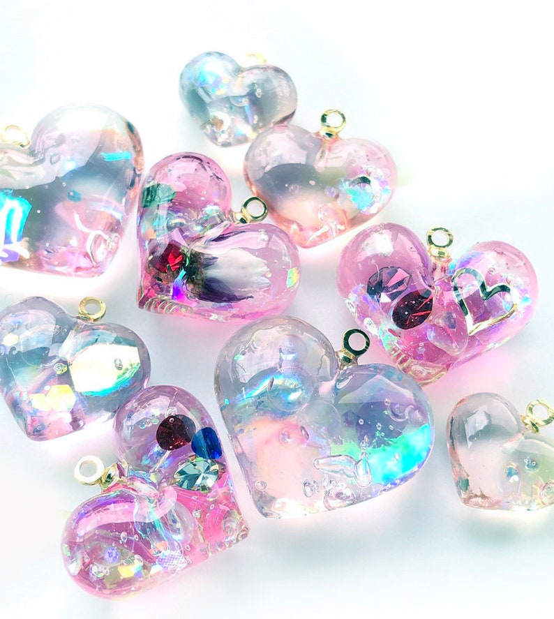May include: A collection of iridescent pink heart-shaped resin charms with gold jump rings. The charms are filled with glitter and other embellishments, creating a sparkly and whimsical effect.
