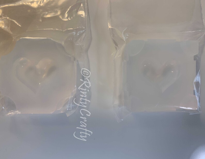 May include: Two clear plastic bags containing white heart-shaped soap bars. The soap bars are in a clear plastic mold. The text "@Rinty Crafty" is visible on the left bag.