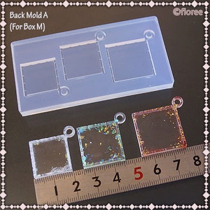 May include: A clear silicone mold with three square cavities for making resin charms. The mold is labeled "Back Mold A (For Box M)". Three resin charms are shown next to the mold, each with a different color and glitter.
