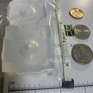 May include: Two clear silicone molds for resin casting, each with a circular indentation. The molds are approximately 1 inch in diameter and are shown next to a ruler and US coins for scale.