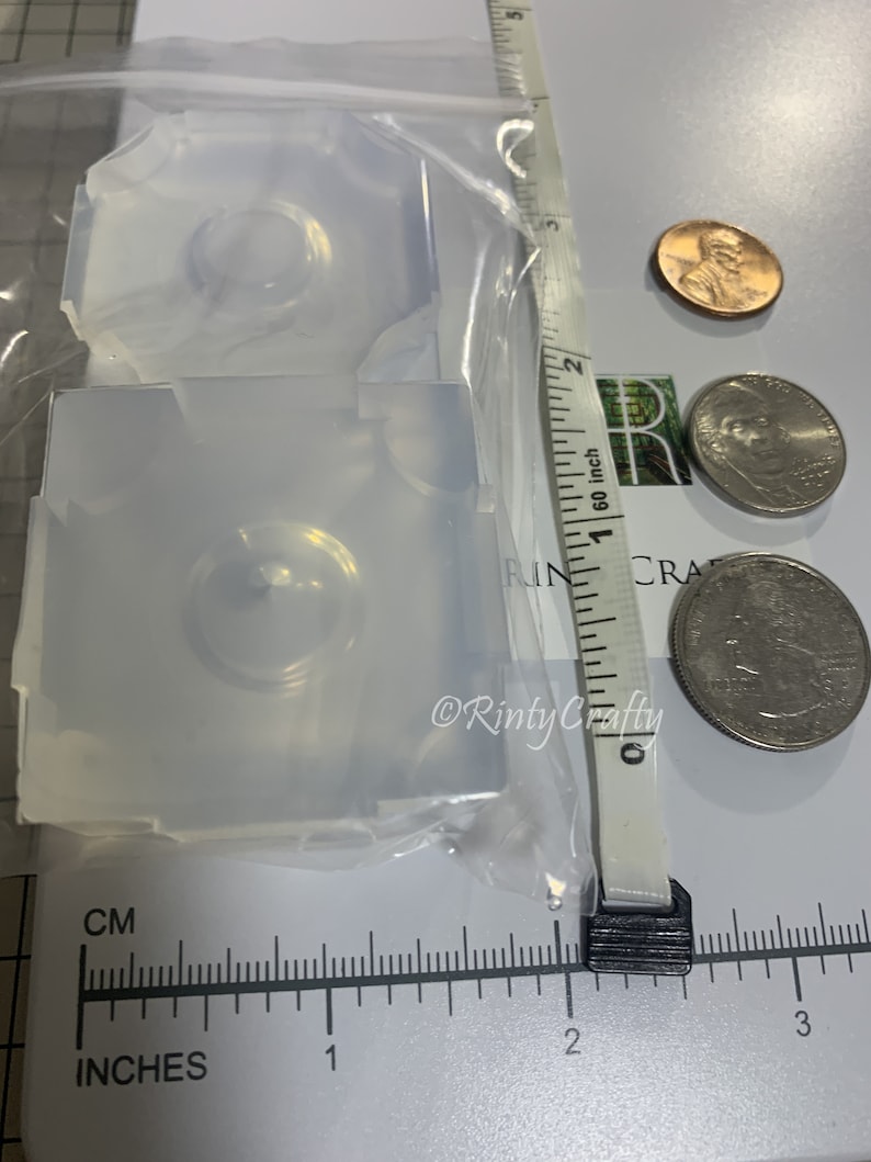 May include: Two clear silicone molds for resin casting, each with a circular indentation. The molds are approximately 1 inch in diameter and are shown next to a ruler and US coins for scale.