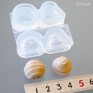 May include: Two clear silicone moulds for making small cake pops. The moulds are divided into two sections, each with a different design. The moulds are shown next to two finished cake pops, one round and one cone-shaped.