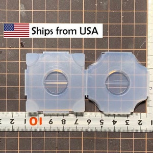 May include: Two clear plastic molds for making resin crafts. Each mold has a square shape with a round opening in the center. The molds are sitting on a gray grid surface.