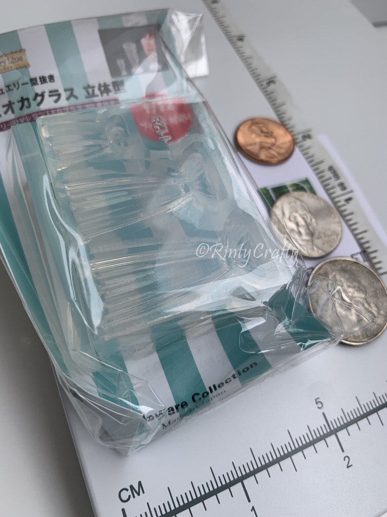 May include: A clear plastic mold for making miniature glass cups. The mold is in a clear plastic bag and is sitting on a white surface with a ruler and coins.