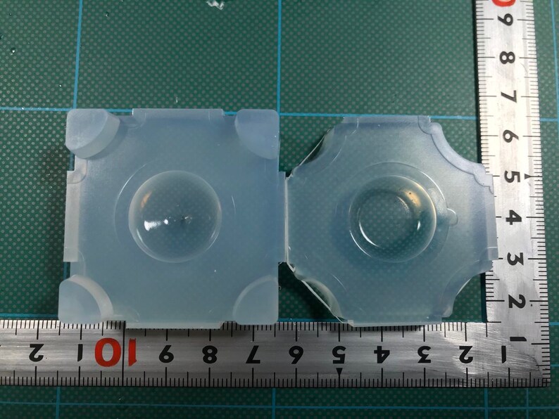 May include: Two clear silicone molds for making resin crafts. The molds are square and round, and are sitting on a green cutting mat with a ruler.
