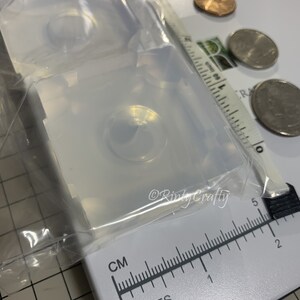 May include: Two clear silicone molds for making resin eyes. The molds are square and have a round indentation for the eye. The molds are sitting on a white surface with a ruler and coins for scale.