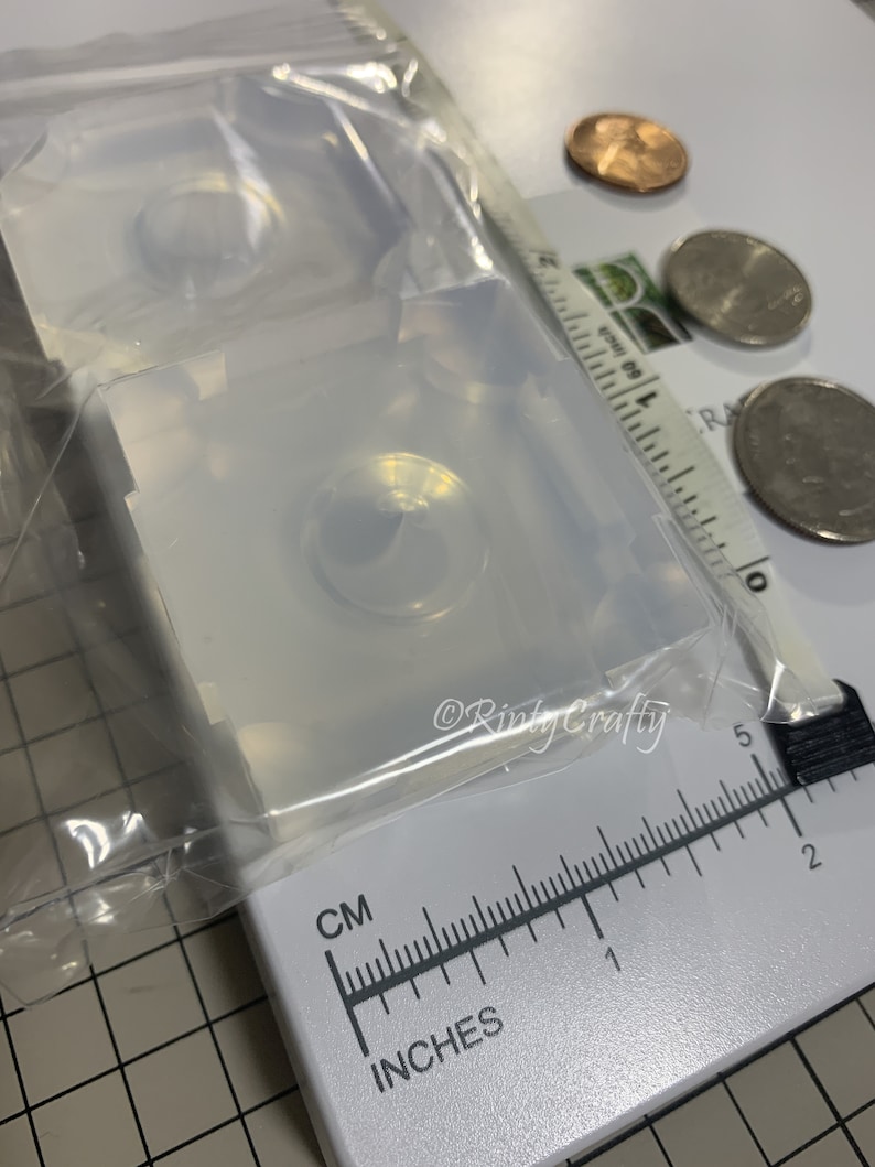 May include: Two clear silicone molds for making resin eyes. The molds are square and have a round indentation for the eye. The molds are sitting on a white surface with a ruler and coins for scale.