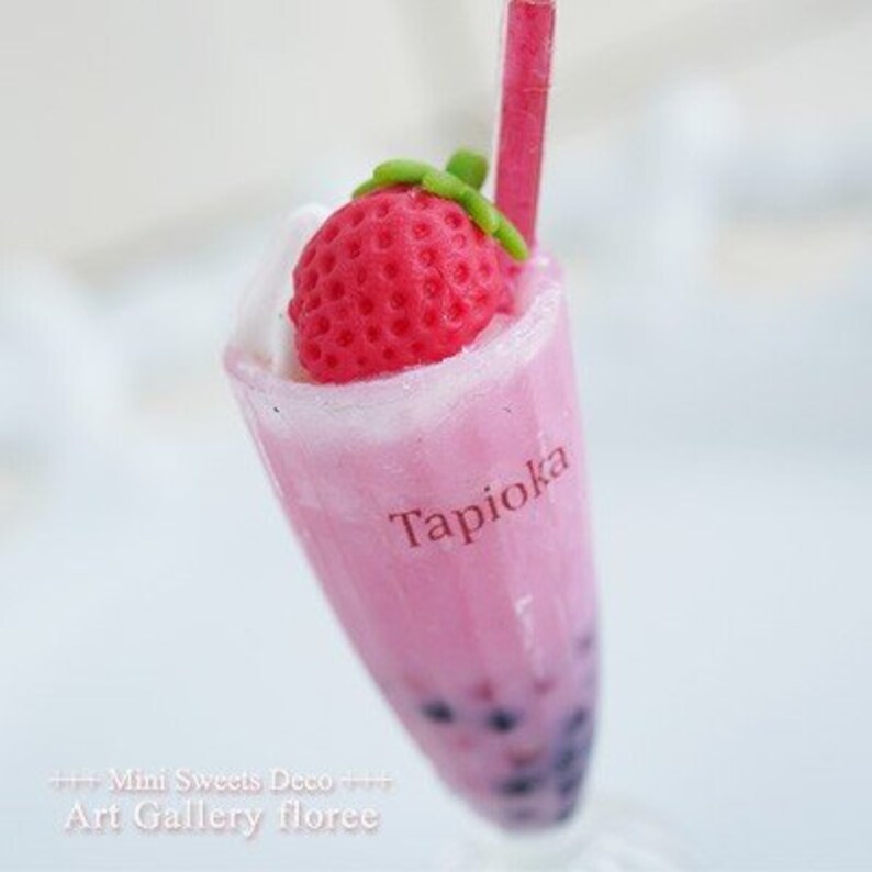 May include: A pink tapioca drink with a strawberry on top and a straw. The drink is in a clear glass with the word "Tapioca" written on the side.