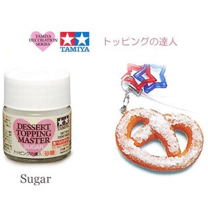 May include: A small white bottle of Tamiya Dessert Topping Master Sugar. The bottle has a pink heart with the words "Dessert Topping Master" and "Sugar" on it. The bottle is labeled in Japanese and English. The bottle is next to a pretzel with powdered sugar on it.