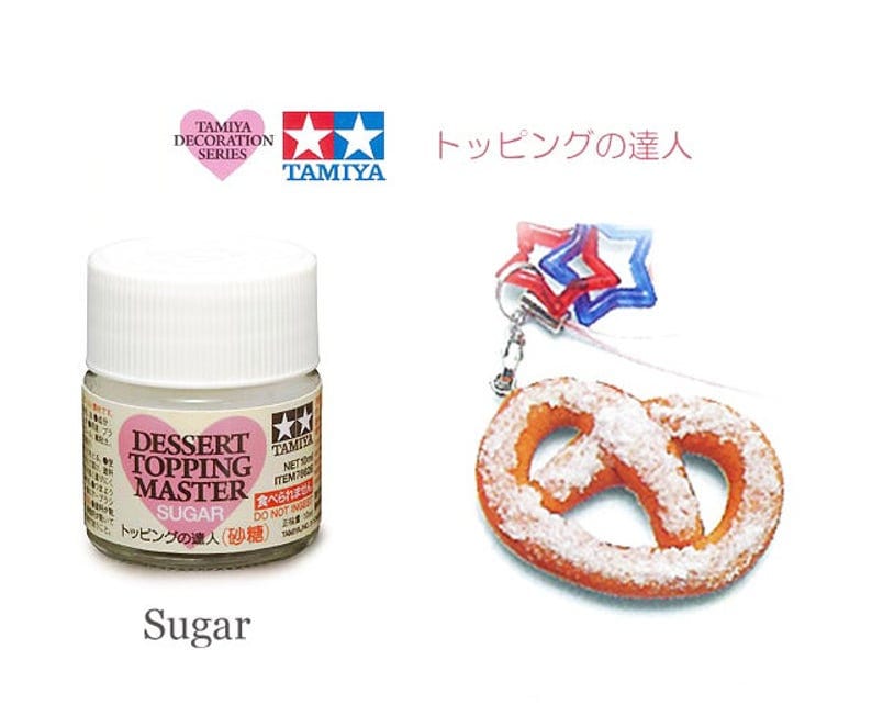 May include: A small white bottle of Tamiya Dessert Topping Master Sugar. The bottle has a pink heart with the words "Dessert Topping Master" and "Sugar" on it. The bottle is labeled in Japanese and English. The bottle is next to a pretzel with powdered sugar on it.