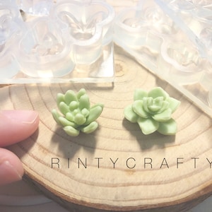 Puede incluir: Dos pequeñas plantas suculentas verdes hechas de resina, sentadas sobre una superficie de madera. Las plantas tienen la forma de una roseta, con múltiples capas de hojas. El fondo muestra un molde de silicona transparente con múltiples cavidades en forma de suculentas.