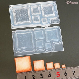 May include: A clear silicone mold with multiple rectangular shapes of varying sizes. The mold is designed for making small bread or pastry shapes. The mold is shown on a gray surface with a ruler and several small, orange-colored bread shapes.