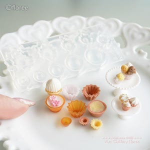 Könnte beinhalten: Eine Sammlung von Miniatur-Desserts und eine Silikonform auf einem weißen, herzförmigen Tablett. Die Desserts umfassen Cupcakes, Törtchen und andere Gebäckstücke in verschiedenen Farben. Die Form ist transparent und weist mehrere kreisförmige und florale Formen auf. Der Text "ofloree" ist sichtbar.