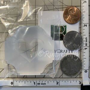 May include: A clear plastic mold with a heart-shaped design. The mold is approximately 1 inch wide and 1 inch tall. The mold is sitting on a white surface with a ruler and coins.