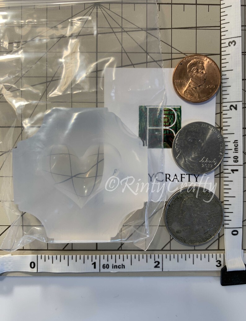 May include: A clear plastic mold with a heart-shaped design. The mold is approximately 1 inch wide and 1 inch tall. The mold is sitting on a white surface with a ruler and coins.
