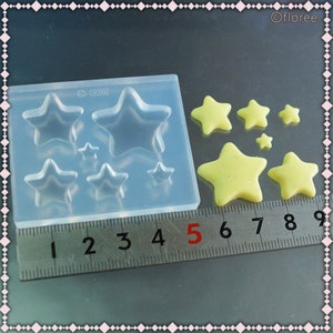 May include: A clear silicone mold with star-shaped cavities of varying sizes, alongside several yellow star-shaped resin pieces. A metal ruler with inch markings is placed next to the mold and stars.