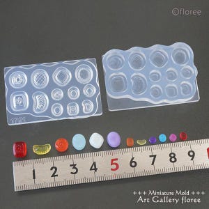 Op de afbeelding: Twee transparante siliconen mallen voor het maken van miniatuurvoedsel. De mallen zijn gevuld met kleine, gedetailleerde vormen, waaronder een bloem, een halve maan, een hart en een ster. De mallen liggen op een wit oppervlak met een liniaal en kleine gekleurde glazen kralen.