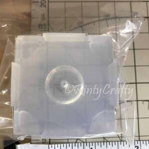 May include: A clear silicone mold for resin casting, featuring a round, recessed area in the center. The mold is approximately 1 inch in diameter.