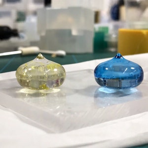 May include: Two small, clear resin ornaments with a rounded, teardrop shape. One ornament is filled with a yellow liquid and the other is filled with a blue liquid.