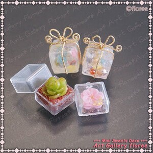 May include: Four miniature gift boxes with clear plastic lids. Two boxes are wrapped with gold ribbon and contain miniature items. One box contains a small green succulent plant and the other contains a pink flower. The boxes are labeled "Mini Sweets Deco Art Gallery floree".