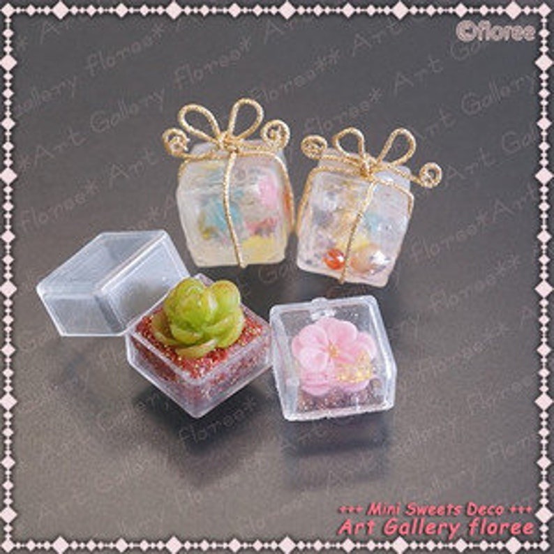 May include: Four miniature gift boxes with clear plastic lids. Two boxes are wrapped with gold ribbon and contain miniature items. One box contains a small green succulent plant and the other contains a pink flower. The boxes are labeled "Mini Sweets Deco Art Gallery floree".