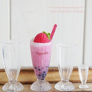 May include: Four miniature glass cups, two with pink tapioca drinks topped with a strawberry, and two empty. The drink cups have the word "Tapioca" printed on them.