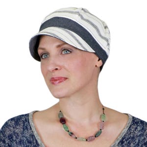 May include: A striped newsboy cap with a dark gray band and brim. The cap features a cream and light gray striped pattern. The woman in the image is wearing a beaded necklace with various colored stones.